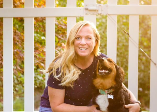 A smiling blonde woman holding a happy brown, black, and white dog with a green collar, in front of a white picket fence and autumn foliage.